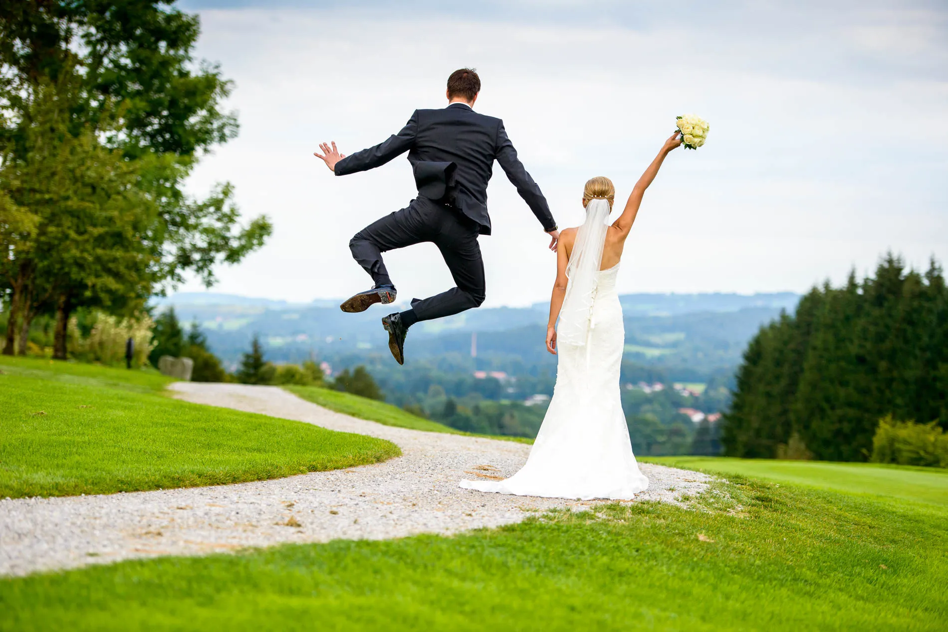 Brautpaar am Hochzeitstag in Waakirchen mit Blick auf die Landschaft, Brautstrauss in der Hand, Bräutigam springt.