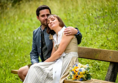Brautpaar auf Bank im Grünen in Bad Kohlgrub, Hochzeitsfotoshooting mit Strauß, traditionell gekleidet.