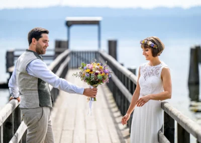 Brautpaar am Ammersee-Pier: Ein Bräutigam reicht der Braut einen Blumenstrauß. Hochzeitsfotografie in Diessen.