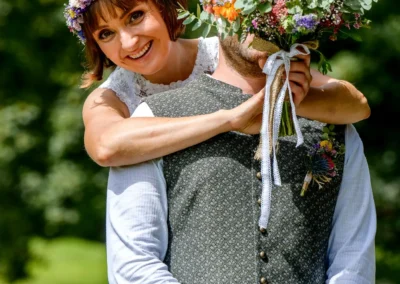 Brautpaar beim Fotoshooting am Ammersee im Schacky Park, Diessen; lachende Braut hinter Bräutigam mit Blumenstrauß.