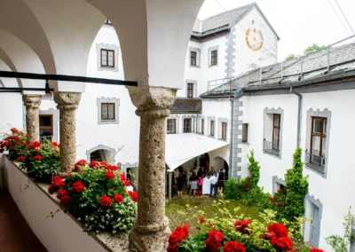 Hochzeit im Innenhof von Schloss Pertenstein, mit Gaesten unter einem Vordach und roten Blumen im Vordergrund.