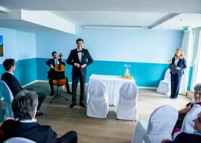 Hochzeitszeremonie in Berg am Starnberger See, Musiker spielt Cello, Rednerin spricht, weiße Stuhlhussen.
