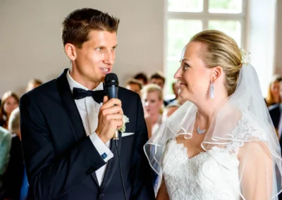 Bräutigam spricht während Hochzeitszeremonie mit Mikrofon, Braut lächelt, Schloss Pertenstein, Chiemgau.