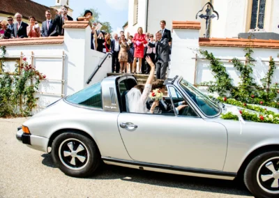 Brautpaar im Oldtimer nach kirchlicher Trauung in Schonstett, Gaeste winken vor Kirche, Hochzeitsreportage Chiemgau.