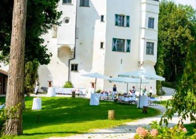 Hochzeitsfeier im Freien bei Schloss Amerang mit gedeckten Tischen und Sonnenschirmen im Garten.