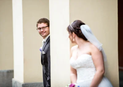Brautpaar bei Fotoshooting im Hofgarten, Muenchen. Hochzeit Reportage zeigt Braut und Bräutigam lachend hinter Saeule.
