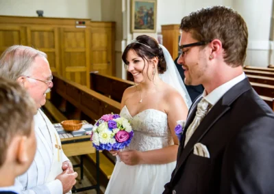 Brautpaar und Priester bei kirchlicher Trauung in Giesing, lächelnde Braut mit Blumenstrauss, Hochzeitsreportage.