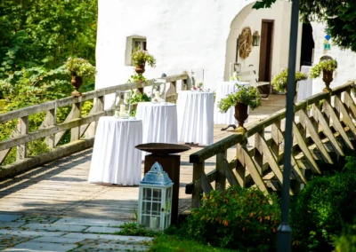 Hochzeitsempfang im Freien auf einer Holzbruecke mit weissen Stehtischen und Blumenarrangements, Schloss Amerang.