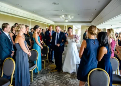 Braut schreitet Arm in Arm mit Begleiter durch Gäste bei Hochzeitsfeier im Hilton Tucherpark München.