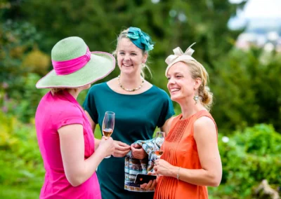 Drei Frauen bei einer Hochzeit im Chiemgau, elegant gekleidet, mit Sektglaesern und Hueten im Freien.