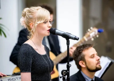 Sängerin bei einer Hochzeit singt vor Mikrofon, Musiker im Hintergrund. Reportage einer Trauung auf Schloss Hohenkammer.