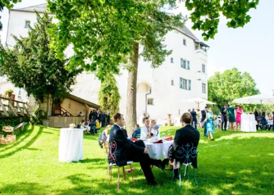 Hochzeitsfeier im Garten von Schloss Amerang, Gaeste in eleganter Kleidung entspannen sich unter Baeumen bei der Trauung.
