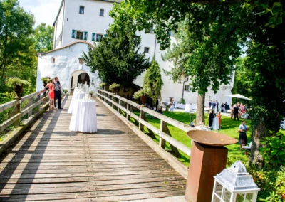 Hochzeitsfeier im Freien bei Schloss Amerang. Gaeste auf Holzsteg und im Park. Hochzeitsreportage mit Schlosskulisse.