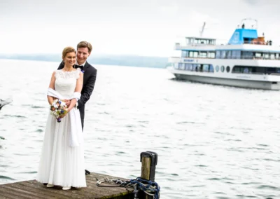 Brautpaar auf Steg am Starnberger See bei Hochzeit, im Hintergrund ein Boot auf dem Wasser. Hochzeitsreportage.