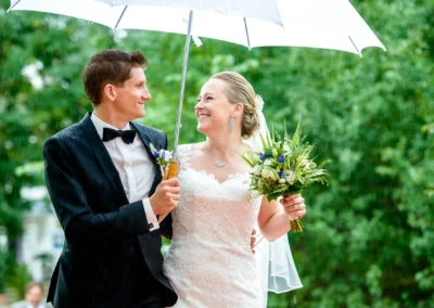 Brautpaar unter Regenschirm bei Hochzeit im Schloss Pertenstein, laechelnd und in eleganter Garderobe mit Blumenstrauss.