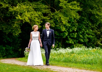Brautpaar bei Hochzeit am Starnberger See; sie spazieren Hand in Hand im Gruen, im Hintergrund Wald und Wiese.
