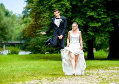 Bräutigam springt neben Braut bei Hochzeitsfeier im Freien, Schloss Pertenstein, festgehaltene Freude und Liebe.