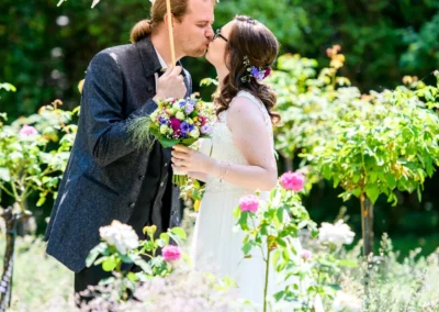 Brautpaar mit Blumenstrauss und Schirm auf Roseninsel im Starnberger See, romantisches Hochzeitsfoto im Garten.