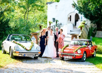 Hochzeitsfoto vor Schloss Amerang mit Brautpaar und Familie, umgeben von zwei dekorierten Oldtimern.
