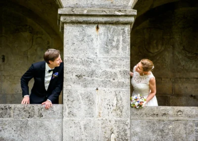 Brautpaar blickt hinter Saeule hervor, romantische Hochzeitsfotografie in Berg am Starnberger See.