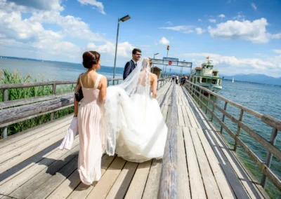 Brautpaar spaziert auf Steg am Chiemsee, Hochzeit im Hotel Aiterbach bei sonnigem Wetter. Romantische Hochzeitsreportage.