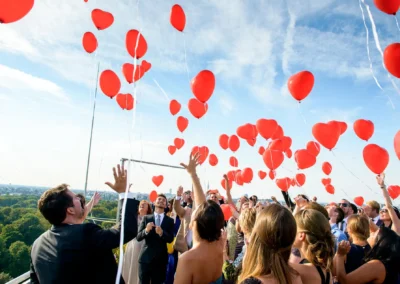 Hochzeitsfeier mit roten Luftballons im Hilton Tucherpark, Muenchen. Fröhliche Trauung mit Gästen unter blauem Himmel.
