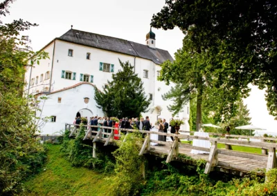 Hochzeitsgesellschaft auf einer Holzbruecke vor Schloss Amerang, umgeben von Baeumen, bei einer Trauung im Chiemgau.