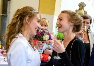 Hochzeitsmoment mit Braut und Gast bei Schloss Aufhausen, Bouquet und Sektgläser, fröhliche Stimmung bei Feier.