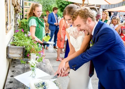 Brautpaar schneidet die Hochzeitstorte im Hasenoehrlhof Geitau an, umgeben von Feiernden in froher Stimmung.