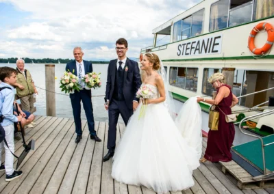 Brautpaar am Steg des Hotels Aiterbach am Chiemsee, begleitet von Gaesten, mit dem Schiff Stefanie im Hintergrund.