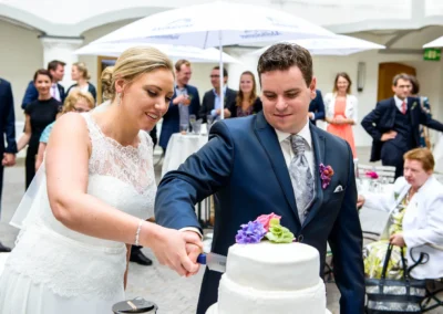 Brautpaar schneidet Hochzeitstorte bei Feier auf Schloss Hohenkammer, umgeben von Gaesten. Hochzeitsfotografie Moment.