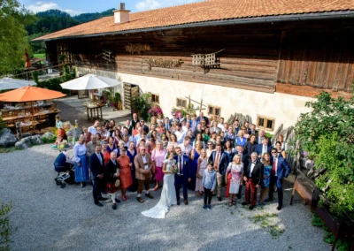 Hochzeitsgesellschaft im Hasenoehrlhof Geitau, Bayrischzell, Gruppenfoto vor traditionellem Gebaeude.
