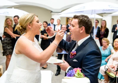 Braut und Braeutigam fuettern sich Hochzeitstorte bei Schloss Hohenkammer-Hochzeit, Gaeste im Hintergrund, Hochzeitsreportage. Preisgekroente Hochzeitsfotografie