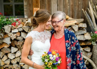 Braut mit Blumenstrauss laechelnd mit aelterer Dame vor Holzhintergrund auf Hochzeit in Geitau am Hasenoehrlhof.