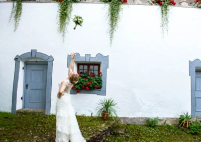 Braut wirft Blumenstrauß vor altem Gebäude. Hochzeit am Schloss Pertenstein, romantische Trauung im Chiemgau.