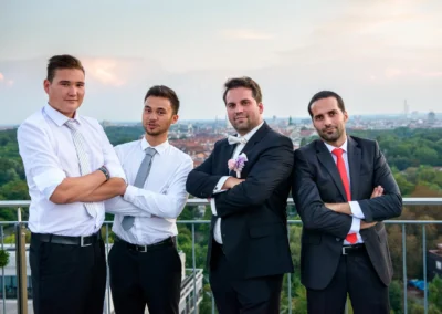 Vier Männer posieren in Anzügen auf einer Hochzeitsfeier in München mit Stadtpanorama im Hintergrund.