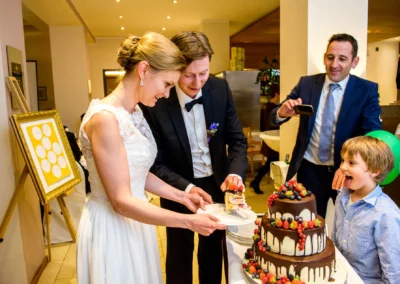 Brautpaar schneidet Hochzeitstorte bei Trauung am Starnberger See, begleitet von Feiernden und Kind mit Luftballons.