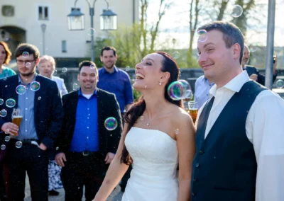 Brautpaar lacht bei Hochzeit in Seefeld, umgeben von Gaesten und Seifenblasen. Hochzeitsreportage im Freien.