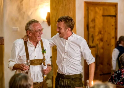 Zwei Maenner in Tracht bei Hochzeit im Hasenoehrlhof, Geitau, lachend und glaesern in der Hand.