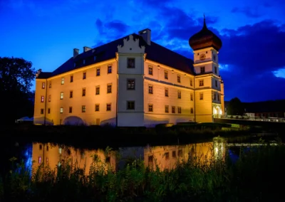 Nachtaufnahme von Schloss Hohenkammer, beleuchtet und malerisch im Wasser reflektiert, ideal fuer Hochzeitsreportage.