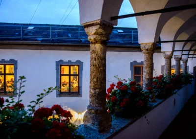 Blick in beleuchtete Fenster bei einer Hochzeitsfeier auf Schloss Pertenstein durch Arkaden mit roten Blumen.