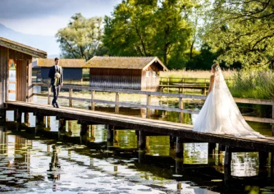 Brautpaar auf Steg am Chiemsee bei Hochzeit im Hotel Aiterbach, umgeben von idyllischer Natur und rustikalen Huetten.