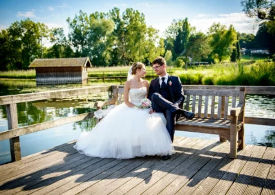 Brautpaar auf Bank am Chiemsee, Hotel Aiterbach, romantische Hochzeit, gruene Landschaft, klarer Himmel.
