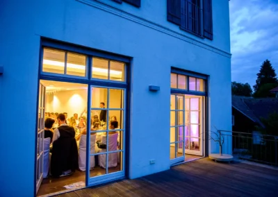 Hochzeitsgesellschaft bei Feier im Hotel Aiterbach, Chiemsee, Abendstimmung zeigt Fenster mit Geselligkeit.
