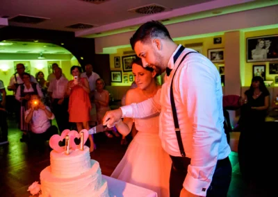 Brautpaar schneidet Hochzeitstorte bei abendlicher Feier in Muenchen an, umgeben von Gaesten.