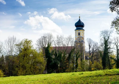 Kirchturm in Bernried am Starnberger See inmitten von Baeumen, gruenem Rasen und blauem Himmel bei einer Hochzeit.