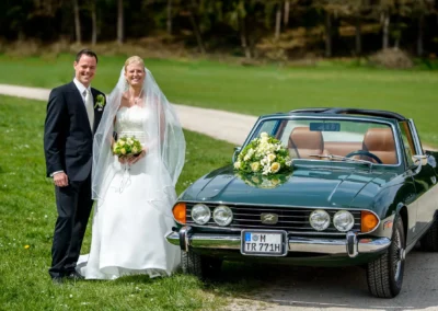 Brautpaar steht vor klassischem Auto bei standesamtlicher Trauung in Oberbayern. Hochzeitsreportage im Gruenen.