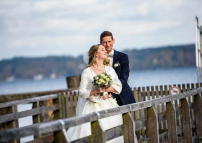 Brautpaar am Steg in Bernried, laecheln verliebt am Starnberger See fuer Hochzeitsfotograf bei einer Hochzeit.