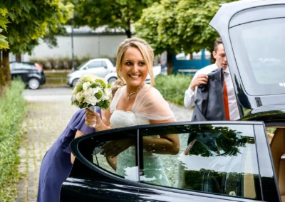 Braut steigt bei kirchlicher Hochzeit in St. Michael, Perlach aus Auto, laechelnd mit Blumenstrauss. Hochzeitsfotografie.