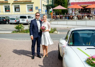 Brautpaar vor Auto bei Hochzeit im Rathaus Tegernsee, strahlender Tag, Hochzeitsfoto in Bayern.